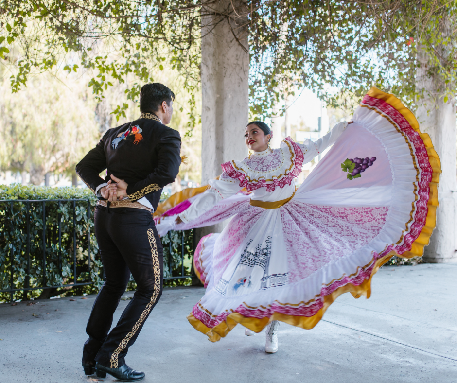 Pareja bailando danza folklórica mexicana; él con traje de charro negro bordado y ella con vestido típico blanco con holanes coloridos, capturados en pleno movimiento durante un zapateado tradicional.