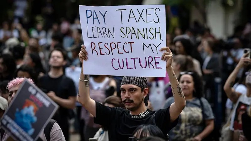 Persona sostiene cartel con el mensaje "Pay taxes. Learn Spanish. Respect my culture" durante una protesta contra la gentrificación en Ciudad de México, rodeada de manifestantes.