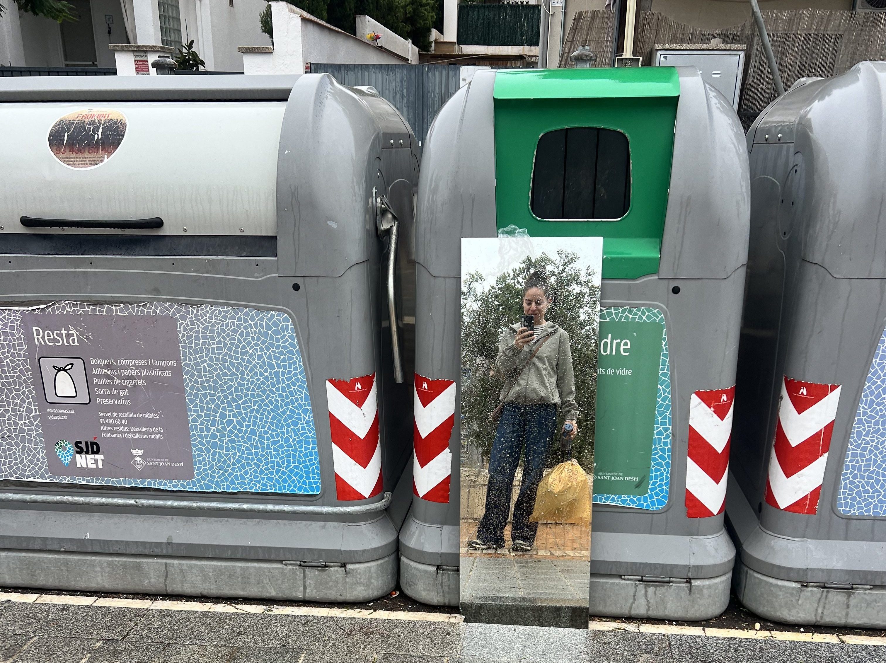 Mujer reflejada en un espejo frente a contenedores de reciclaje, metáfora sobre soltar el exceso de trabajo y mirarse con nuevos ojos.