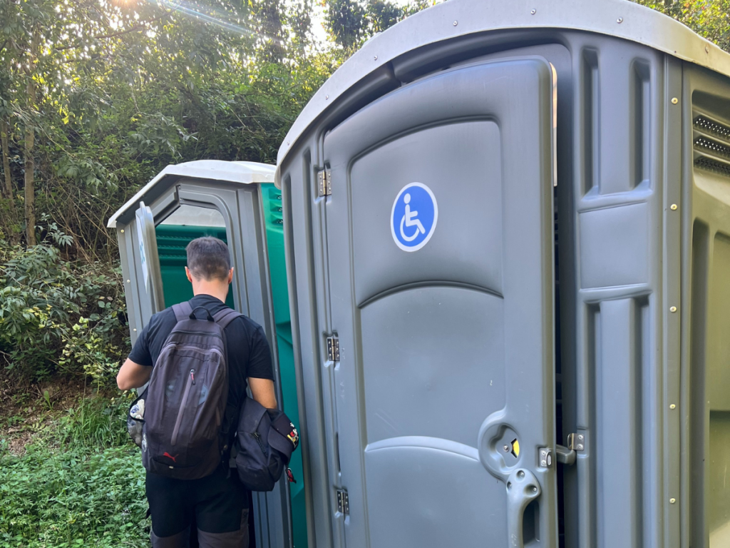 Persona con mochila frente a un baño portátil accesible en medio del bosque, símbolo de la buena infraestructura en rutas de senderismo