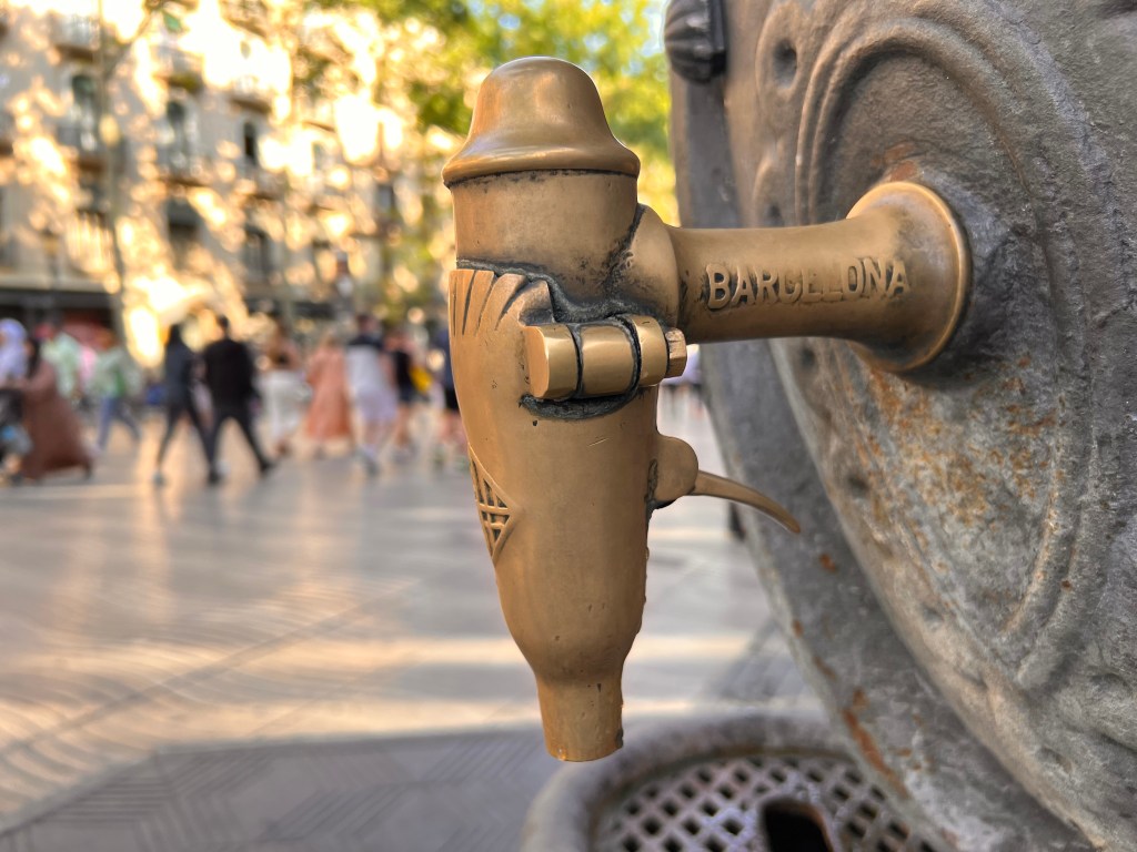 Fuente pública de agua en una calle de Barcelona con personas caminando al fondo