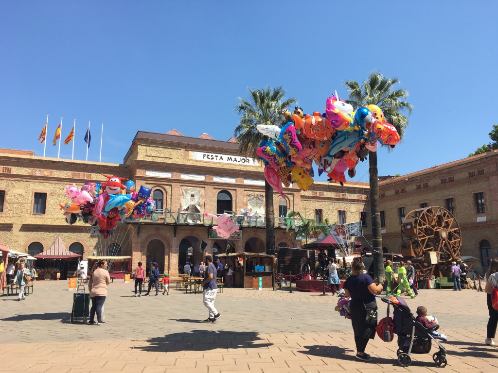 Plaza de Nou Barris en Barcelona con globos de colores y personas caminando durante una fiesta popular en la ciudad